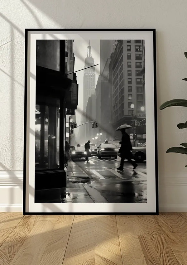 Een zwart-wit ingelijste foto van een regenachtig straatbeeld in de stad, met voetgangers met parasols, auto's en hoge gebouwen, waaronder het Empire State Building op de achtergrond. Dit prachtige Rainy Day Schilderij van CollageDepot leunt tegen een muur op een houten vloer, klaar om te worden opgehangen met een magnetisch ophangsysteem.,Zwart