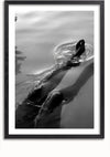 Een zwart-witfoto, die doet denken aan een zwart-wit schilderij, toont een persoon die in kalm water zwemt. Alleen de benen en voeten van de persoon zijn zichtbaar als ze door het oppervlak breken, waardoor er kleine rimpelingen omheen ontstaan. De afbeelding is elegant ingelijst in een eenvoudig zwart frame, perfect voor wanddecoratie. Dit is het "Relaxed baden schilderij" van CollageDepot.
