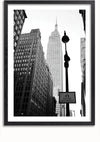 Zwart-witfoto van het Empire State Building, gezien tussen hoge stadsgebouwen. Op de voorgrond zijn een straatlantaarn en een bord met de tekst "Bus Lanes Photo Enforced" zichtbaar. Dit boeiende Streets of New York schilderij van CollageDepot is ingelijst met een zwarte lijst, perfect voor wanddecoratie met een magnetisch ophangsysteem.