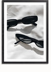 Op een ingelijste foto staan twee zwarte zonnebrillen op een wit oppervlak. Het bovenste paar is rechthoekig en het onderste paar heeft de vorm van een kattenoog. Schaduwen van de zonnebril zijn zichtbaar op het oppervlak, waardoor het een perfecte wanddecoratie is voor elke kamer. Maak kennis met het Twee Zwarte Zonnebrillen Schilderij van CollageDepot.,Zwart-Met,Lichtbruin-Met,showOne,Met